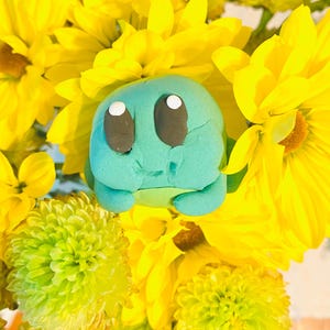 May include: A teal-colored, cartoon-style figure with large black eyes and a smiling expression, surrounded by bright yellow flowers. The figure has a rounded shape and is set against a backdrop of vibrant yellow petals and green foliage.