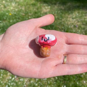May include: A small, hand-painted mushroom with a red and white cap and a brown stem. The mushroom is painted with the word "luv" on the cap.