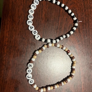 May include: Two beaded bracelets. One bracelet features black and white beads with the word "SERGEANT" spelled out in white letter beads. The other bracelet has brown, tan, and white beads with the word "KOJO" spelled out in white letter beads.