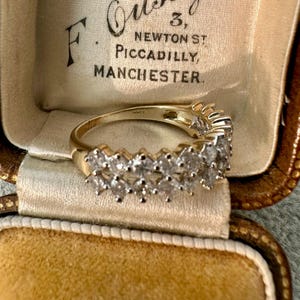 May include: A gold ring with multiple clear gemstones set in a row. The ring is displayed inside a vintage jewelry box with text that reads "F. Ousey, 3, Newton St, Piccadilly, Manchester."