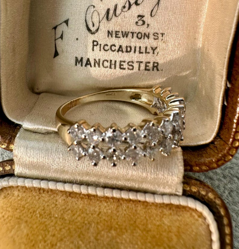 May include: A gold ring with multiple clear gemstones set in a row. The ring is displayed inside a vintage jewelry box with text that reads "F. Ousey, 3, Newton St, Piccadilly, Manchester."