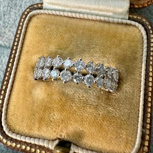 May include: A gold ring with two rows of clear, sparkling gemstones. The ring is set in a gold band and is displayed in a vintage jewelry box with a tan interior.