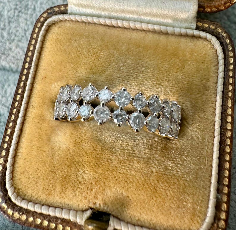 May include: A gold ring with two rows of clear, sparkling gemstones. The ring is set in a gold band and is displayed in a vintage jewelry box with a tan interior.