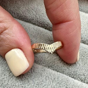 May include: A gold-colored chevron ring with a textured surface. The ring has a V-shape design and is held between two fingers. The ring's surface has a series of vertical lines.