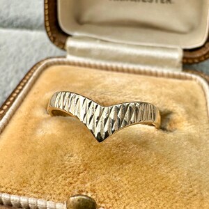 May include: A gold-colored chevron ring with a textured surface. The ring is displayed inside a vintage jewelry box with a tan interior. The ring's design features a V-shape with a series of diagonal lines.