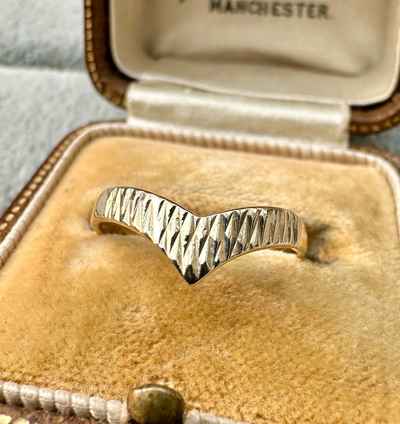 May include: A gold-colored chevron ring with a textured surface. The ring is displayed inside a vintage jewelry box with a tan interior. The ring's design features a V-shape with a series of diagonal lines.