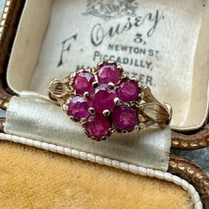 以下が含まれることがあります： 7つの丸い鮮やかな赤い宝石のクラスターが特徴のヴィンテージゴールドリング。 リングは花柄のデザインで、バンドには複雑なディテールが施されています。 リングはヴィンテージのジュエリーボックスの中に展示されています。