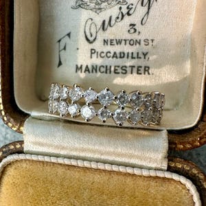 May include: A sparkling diamond ring with a double-row design. The ring features numerous round-cut diamonds set in a band. The ring is displayed inside a vintage jewelry box with text that reads "Ousey, 3, Newton St, Piccadilly, Manchester."