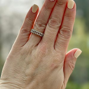 May include: A diamond ring with a double-row design is displayed on a hand with manicured nails. The ring features multiple small, clear diamonds set closely together. The nails are painted in shades of beige and pink.