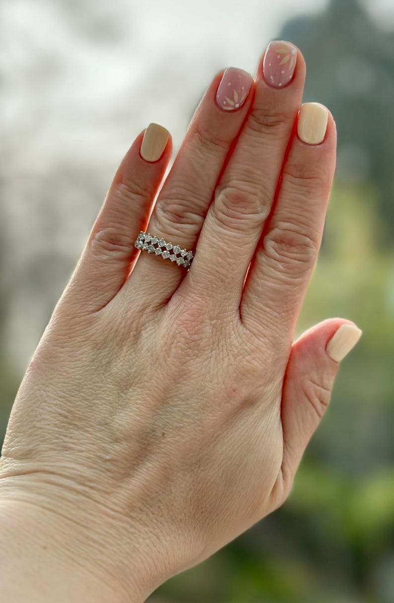 May include: A diamond ring with a double-row design is displayed on a hand with manicured nails. The ring features multiple small, clear diamonds set closely together. The nails are painted in shades of beige and pink.