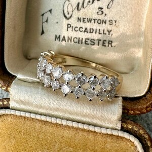 May include: A gold ring with multiple clear gemstones set in a row. The ring is displayed inside a vintage jewelry box with text that reads "F. Ousby, 3, Newton St, Piccadilly, Manchester."