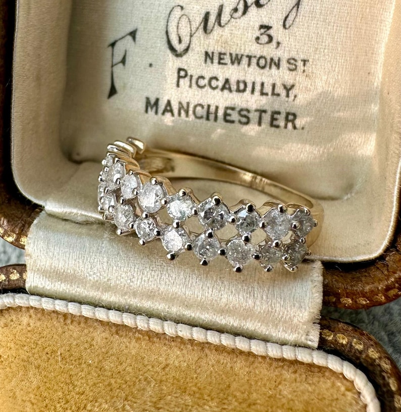 May include: A gold ring with multiple clear gemstones set in a row. The ring is displayed inside a vintage jewelry box with text that reads "F. Ousby, 3, Newton St, Piccadilly, Manchester."