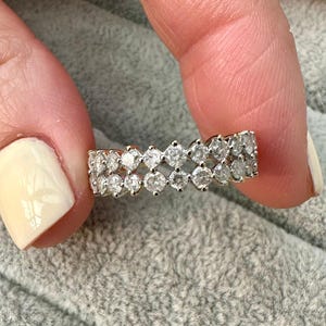 May include: A gold ring with two rows of clear, round-cut gemstones. The ring is held between two fingers against a soft, gray background. The gemstones are set closely together, creating a sparkling effect.