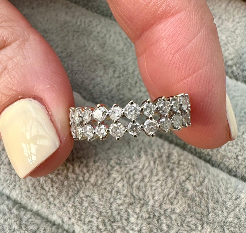 May include: A gold ring with two rows of clear, round-cut gemstones. The ring is held between two fingers against a soft, gray background. The gemstones are set closely together, creating a sparkling effect.