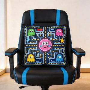 Puede incluir: Cojín cuadrado negro con un diseño pixelado de una cara sonriente rosa, fantasmas, cerezas y una estrella. El cojín está sobre una silla de juego negra y azul. El diseño recuerda a un juego de arcade clásico.