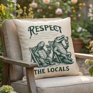 May include: A square off-white throw cushion featuring a dark green graphic of a turtle and an alligator sitting in beach chairs, each holding a tropical drink. The text "RESPECT" is printed above and "THE LOCALS" is printed below the graphic.