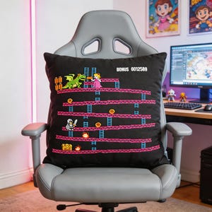 May include: Black square pillow featuring a pixelated video game scene with a dragon, princess, and knight. The design includes pink platforms, blue ladders, and the text "BONUS 0012589". The pillow is on a gray gaming chair.