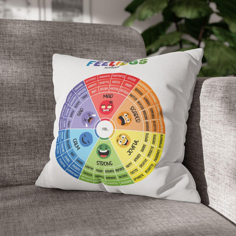 May include: A white square throw pillow featuring a colorful feelings wheel graphic. The design includes six primary emotional categories labeled Mad, Scared, Joyful, Strong, Calm, and Sad, each with sub-emotions and corresponding emoji faces.