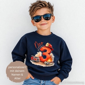 Könnte beinhalten: Ein dunkelblaues Langarm-Sweatshirt mit einem Grafikdruck, der eine große rote Zahl Drei, ein rotes Feuerwehrauto, einen Feuerwehrschlauch und einen gelben Feuerwehrhelm zeigt. Der Name Neo ist in Orange über der Zahl aufgedruckt.