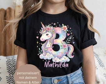 Puede incluir: Una camiseta negra de manga corta con un gráfico colorido de un unicornio entrelazado con el número 3, decorado con flores, estrellas y nubes. El nombre Mathilda está impreso en rosa debajo del gráfico. La camiseta tiene cuello redondo.
