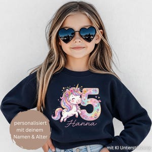 Könnte beinhalten: Ein dunkelblaues Langarm-Sweatshirt mit einer Grafik eines weißen Einhorns mit bunter Mähne, einem Blumenkranz und der mit Blumen verzierten Zahl 5. Der Name Hanna steht darunter in Schreibschrift, ergänzt durch einen runden Textaufkleber.