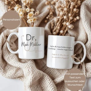 Può includere: Due tazze in ceramica bianca con testo nero e un disegno di stetoscopio. Una tazza recita "Dr. Max Müller" con una linea di battito cardiaco. L'altra tazza ha testo tedesco. Le tazze sono su una superficie beige a maglia.