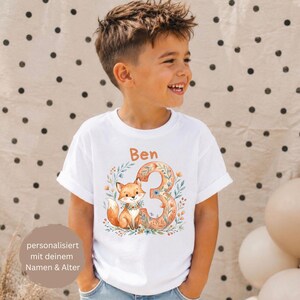 Könnte beinhalten: Ein weißes T-Shirt mit kurzen Ärmeln und einem Aufdruck, der einen Fuchs neben einer großen Zahl 3 zeigt, umgeben von floralen Elementen. Der Name Ben ist in Orange darüber gedruckt. Das Design verwendet orange, blaue und grüne Farbtöne.