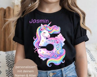 Puede incluir: Una camiseta negra con un gráfico colorido de un unicornio con forma de número cinco, decorado con flores, estrellas y una luna creciente. El nombre Jasmin está impreso en morado sobre el gráfico, junto a una superposición de texto circular.