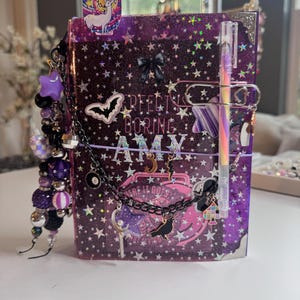 May include: A purple glittery journal with star patterns, featuring the text "PERFECT IS BORING" and "AMY". It is decorated with a black bow, stickers of a bat and a raven, a chain with charms, and a pen attached by a large silver paperclip.