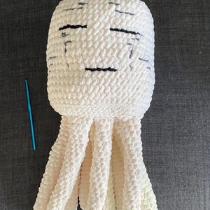 May include: A handmade, cream-coloured crochet jellyfish plush toy with black stitched facial features. The jellyfish has multiple cream-coloured tentacles and a blue crochet hook is visible. The plush is made of a soft, textured yarn.