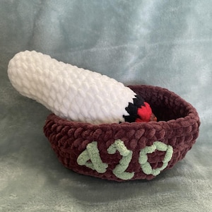 May include: A handmade crochet piece. A large, white, cylindrical object with a black and red tip sits in a burgundy bowl. The bowl has the number "420" in green yarn stitched on the side.