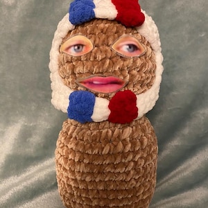 May include: A plush, brown, handmade doll with a white hood and a blue and red trim. The doll has eyes and a mouth attached to the face. The doll is a unique, whimsical creation.