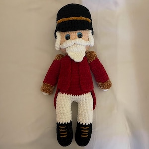 May include: Crocheted nutcracker doll in a classic soldier uniform. The doll features a black hat with a gold band, a red jacket with gold trim, white pants, and black boots. The face has blue eyes and a white beard.