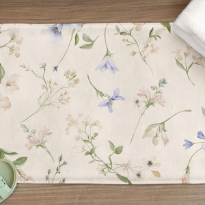 Può includere: Tappetino da bagno rettangolare con sfondo color crema e motivo floreale ad acquerello. Il design presenta delicati fiori blu, rosa e bianchi con foglie verdi. Un asciugamano bianco piegato è appoggiato sul tappetino. Sono visibili sandali verde chiaro.