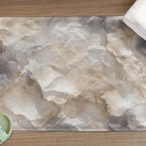 May include: A rectangular bath mat with a stone-like pattern in shades of gray, beige, and white. A white towel is folded on the right side. Light green sandals are on the lower left corner. The mat has a textured appearance.