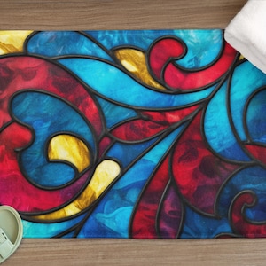 May include: A rectangular bath mat with a stained glass design in shades of blue, red, and yellow. The mat has a black border and is next to a pair of green sandals and a white towel.