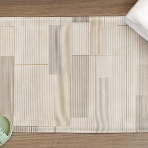 Op de afbeelding: Rechthoekige badmat met een geometrisch patroon in beige, grijs en bruin. Een witte opgevouwen handdoek ligt op de mat. Lichtgroene sandalen liggen in de buurt van de mat op een houten oppervlak.