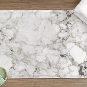 May include: A rectangular bath mat with a marble pattern in shades of white, gray, and black. A folded white towel rests on the mat. Light green sandals are at the bottom left corner. The mat measures approximately 30 inches long.