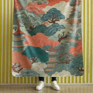 Könnte beinhalten: Eine Decke mit einem japanisch inspirierten Landschaftsdesign. Das Design umfasst Pagoden, Bäume und Wolken in Türkis, Koralle und Creme. Die Decke wird vor einer gelb-weiß gestreiften Wand gehalten.