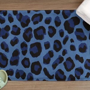 May include: Blue and black leopard print bath mat. The rectangular mat features a repeating pattern of dark blue and black leopard spots on a light blue background. A white towel and light green sandals are also visible.