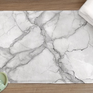 May include: A rectangular bath mat with a white marble design, featuring gray veining. A folded white towel rests on the mat's edge. Light green sandals are placed near the mat on a wooden surface. The mat is approximately 76 cm long.