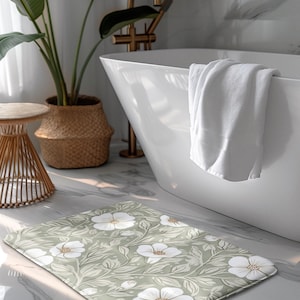 May include: A rectangular bath mat featuring a floral design of white flowers and green leaves on a pale green backdrop. The mat is positioned on a marble floor in a bathroom, near a white bathtub.