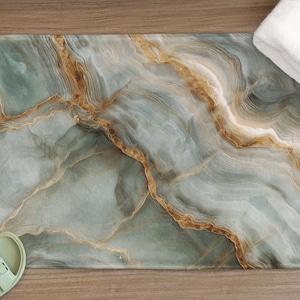 May include: A rectangular bath mat with a marble design in shades of teal, grey, and gold. The mat has rounded corners and is next to a pair of light green sandals and a folded white towel. The bath mat is on a wooden surface.