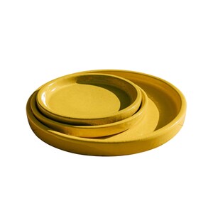 Könnte beinhalten: Ein Stapel aus drei gelben Keramikplatten in verschiedenen Größen. Die Teller sind ineinander verschachtelt, wobei der kleinste Teller oben und der größte unten liegt. Die Teller haben eine glatte, glänzende Oberfläche und ein schlichtes, modernes Design.