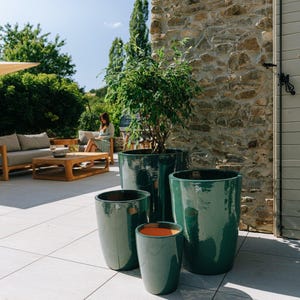 Peut inclure: Ensemble de quatre pots de fleurs en céramique vert émeraude brillant de différentes tailles. Un pot a un intérieur orange. Les pots sont disposés sur un patio carrelé, avec un mur en pierre et de la verdure en arrière-plan.
