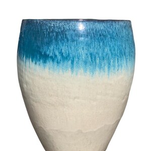 Könnte beinhalten: Eine Keramikvase in konisch zulaufender zylindrischer Form mit einer glatten, cremeweißen Basis, die am oberen Rand in eine leuchtend blaue, verlaufende Glasur übergeht. Die Oberfläche weist eine dezente, handgefertigte Textur mit Farbnuancen auf.