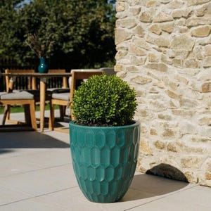 Könnte beinhalten: Ein türkisfarbener Keramik-Blumentopf mit geometrischem Muster enthält einen beschnittenen grünen Strauch. Die Außenszene umfasst eine Steinmauer, Gartenmöbel und einen weißen Sonnenschirm. Der Topf ist ein dekoratives Element für Gärten und Terrassen.