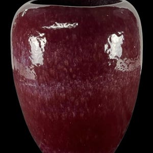 Könnte beinhalten: Eine tief burgunderfarbene Vase mit glänzendem Finish. Die Vase hat einen abgerundeten Körper und eine weite Öffnung. Die Oberfläche hat eine gesprenkelte Textur und die Gesamtform ist leicht oval. Der Hintergrund ist schwarz.