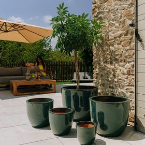 Könnte beinhalten: Eine Sammlung dunkelgrüner, glasierter Keramik-Pflanzgefäße in verschiedenen Größen. Die Pflanzgefäße stehen auf einer hell gefliesten Terrasse, mit einem Baum im größten Topf. Die Szene spielt im Freien unter sonnigem Himmel.