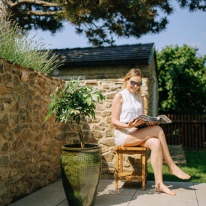 Könnte beinhalten: Eine Frau mit Sonnenbrille und weißem Oberteil ohne Ärmel liest im Freien eine Zeitschrift. Ein großer grüner Keramiktopf mit einer Pflanze steht daneben. Eine Steinmauer und Bäume sind im Hintergrund.
